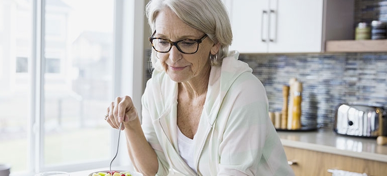 Mature woman eating in kitchen while reading on a tablet